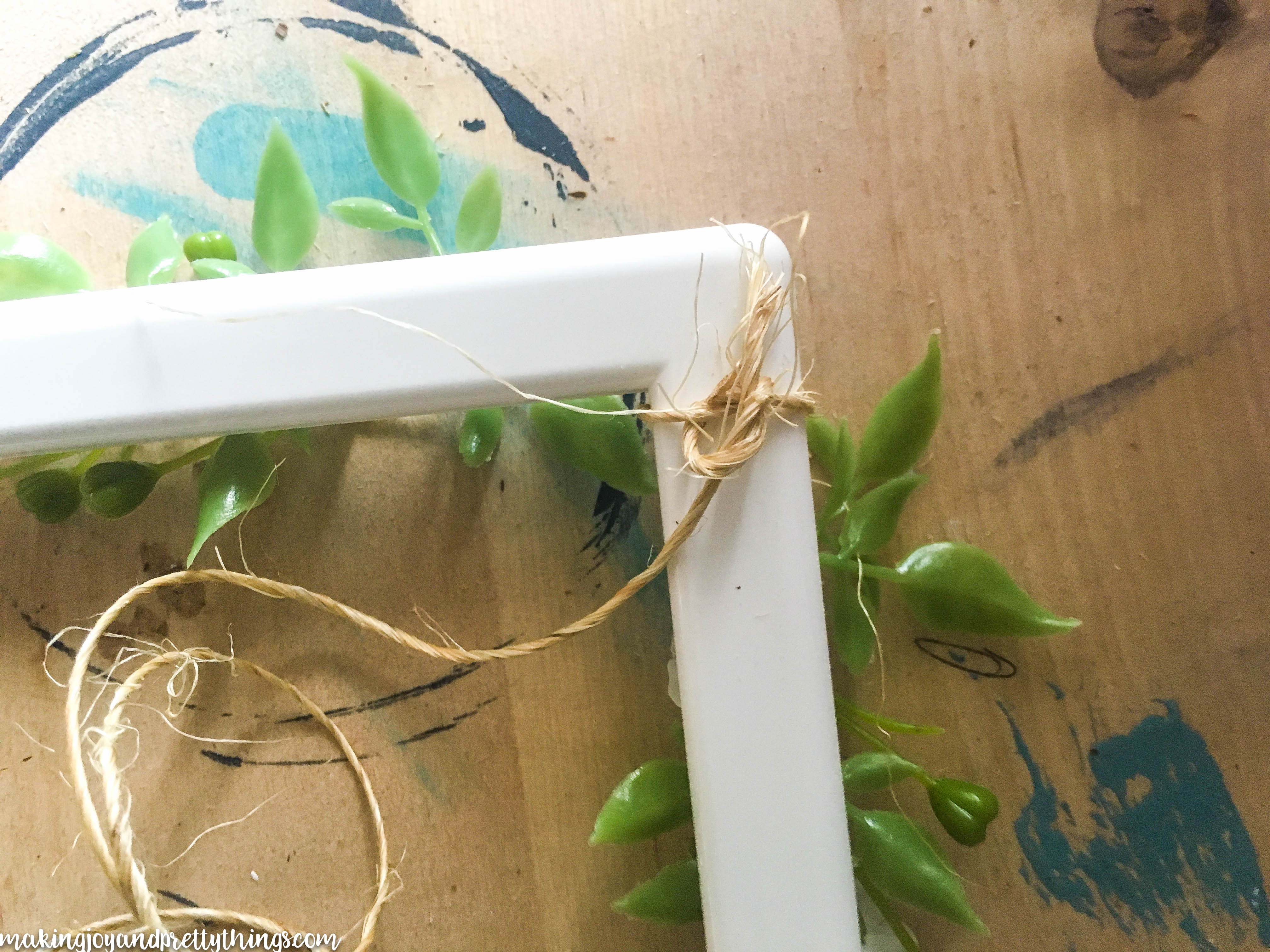 A long piece of craft twine is tied in a knot around the corner of a white picture frame. Faux greenery sprigs are visible, glued to the front side of the frame.