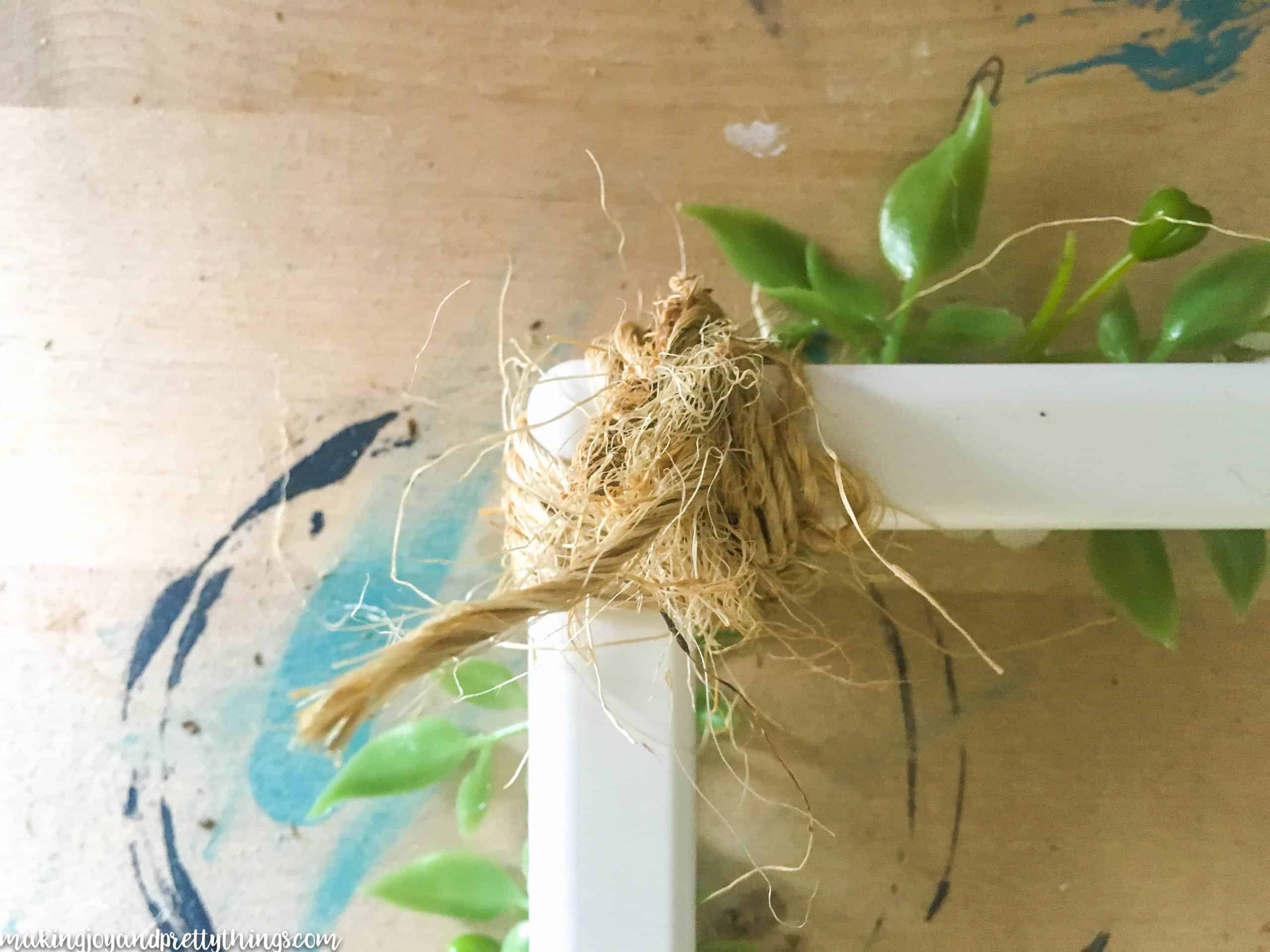 A string of crafting twine is wrapped around the corner of a white picture frame and tied off with a knot. There are sprigs of faux greenery glued to the front side of the picture frame.