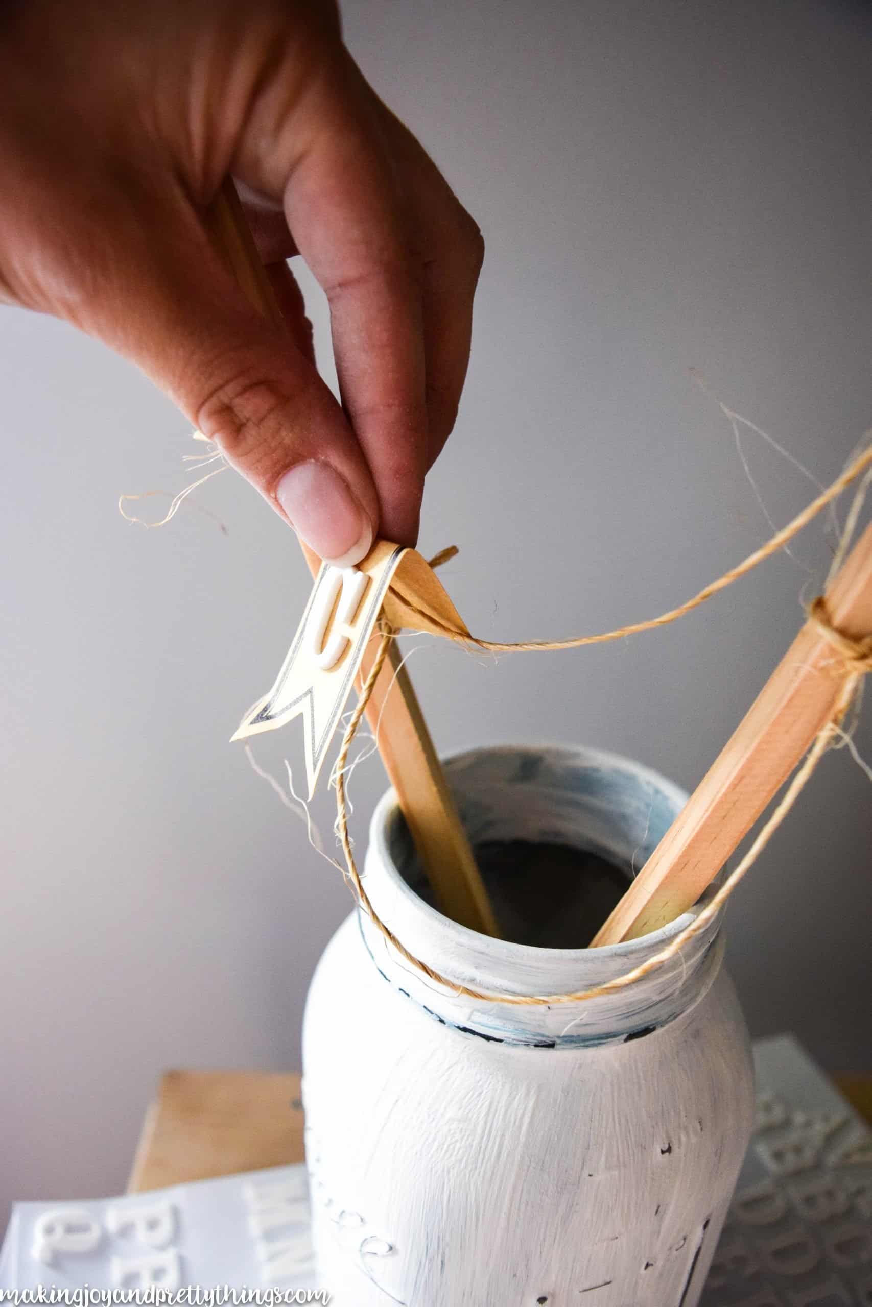 White painted distressed mason jar with dowel rods in it and twine strung across to hang lettering 