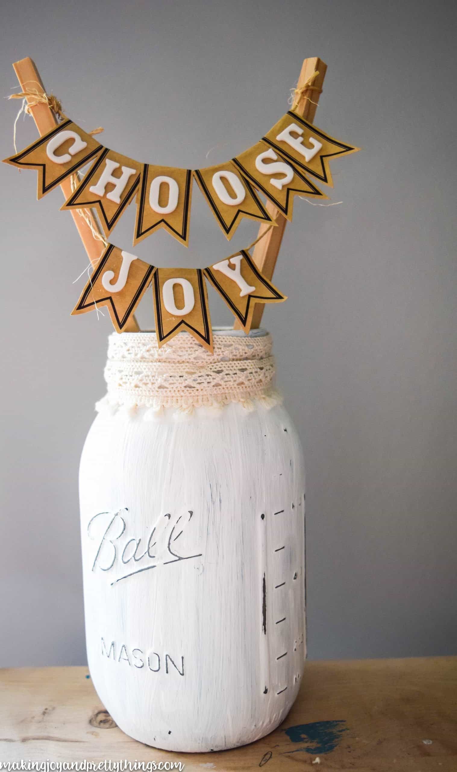 A white painted mason jar with a lace trim around the top. Two thin sticks hold a small paper banner that reads "choose joy"