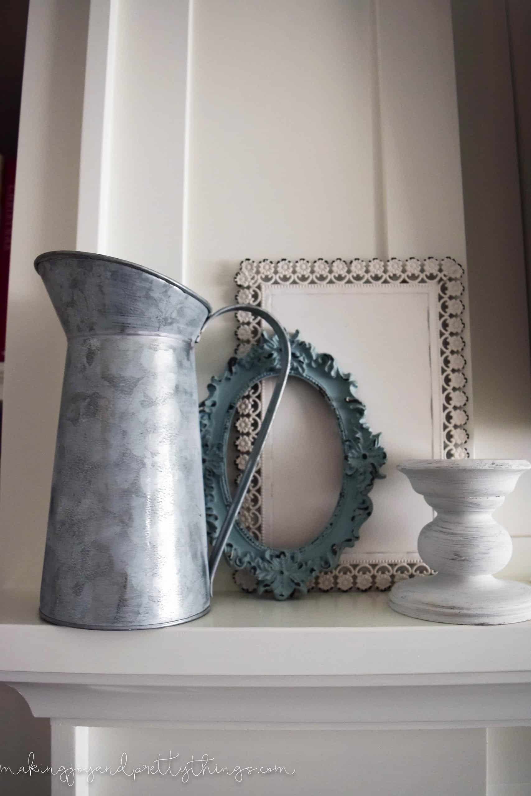 A galvanized metal pail stands next to two empty picture frames and a rustic white painted candle holder on a white fireplace mantle.