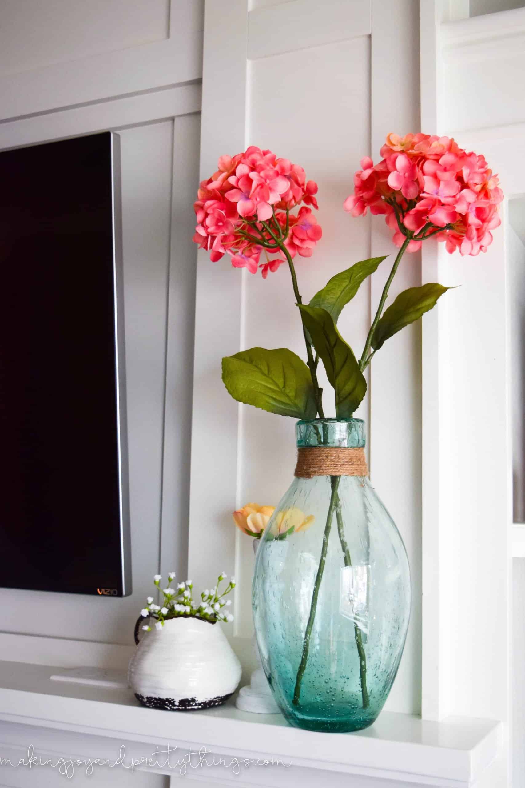 A blue seaglass vase filled with two faux stems of bright pink hydrangea flowers sits on a white mantle.