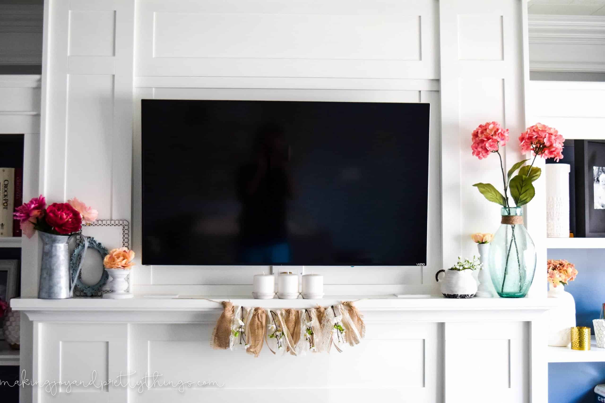 A look at the completed summer mantle with vases of faux summery flowers and other farmhouse decor pieces. A large flatscreen TV is mounted in the center of the white mantle.