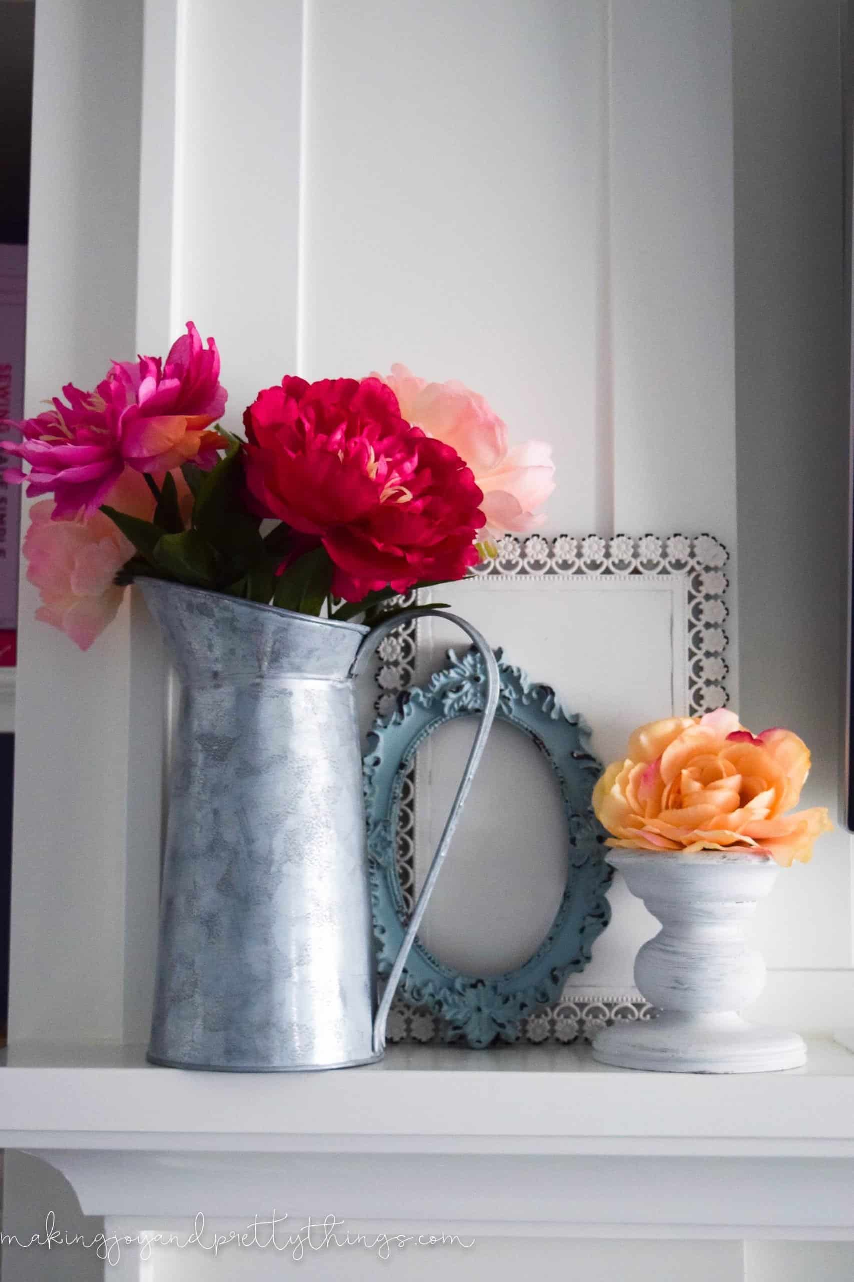 A galvanized metal vase filled with bright pink and red faux stems of flowers sits on a white mantle, next to a candle holder and empty painted picture frames.