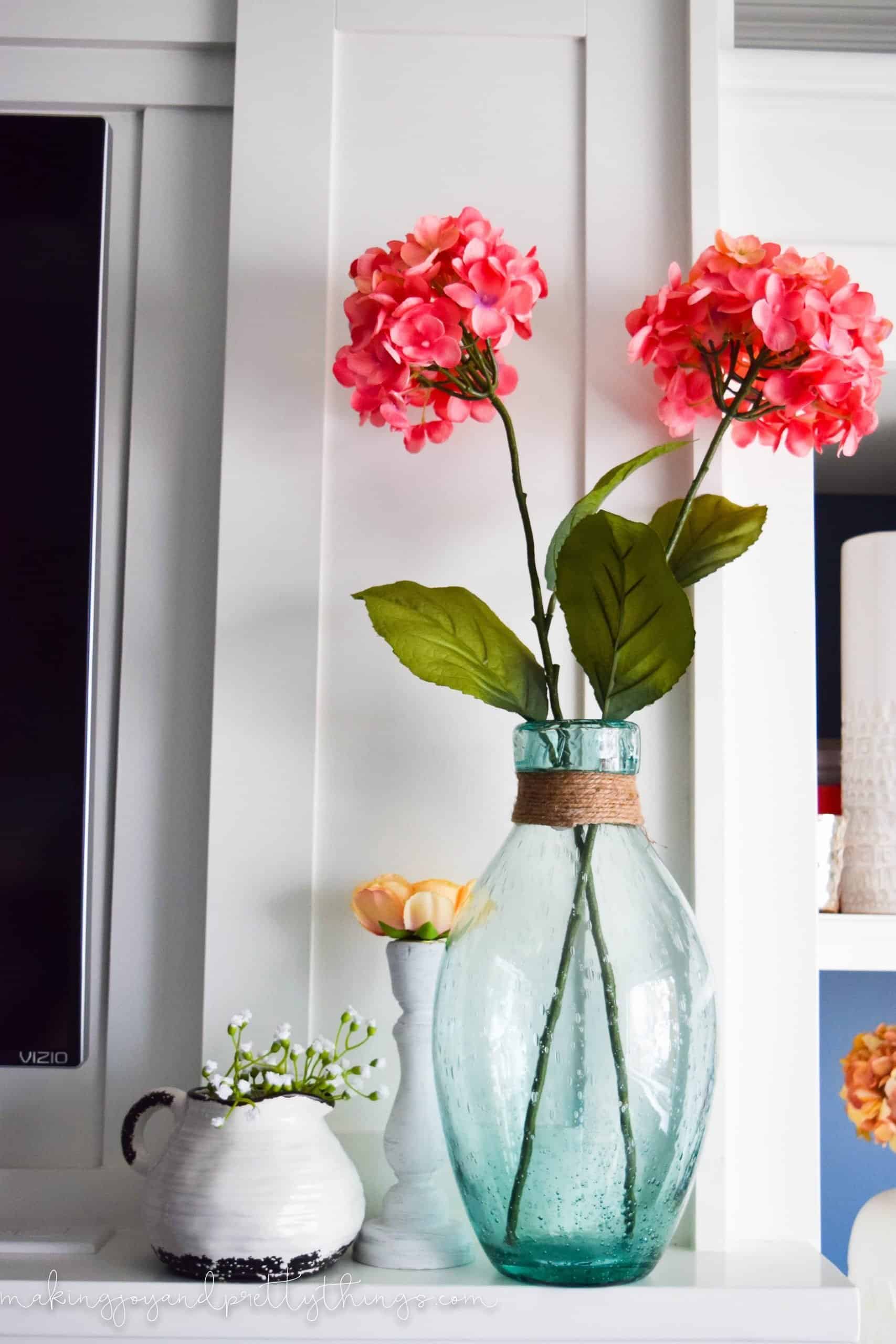 Two stems of bright pink hydrangea blooms in a blue seaglass vase. The vase sits on a mantle next to a small white ceramic jar and rustic candle holder.