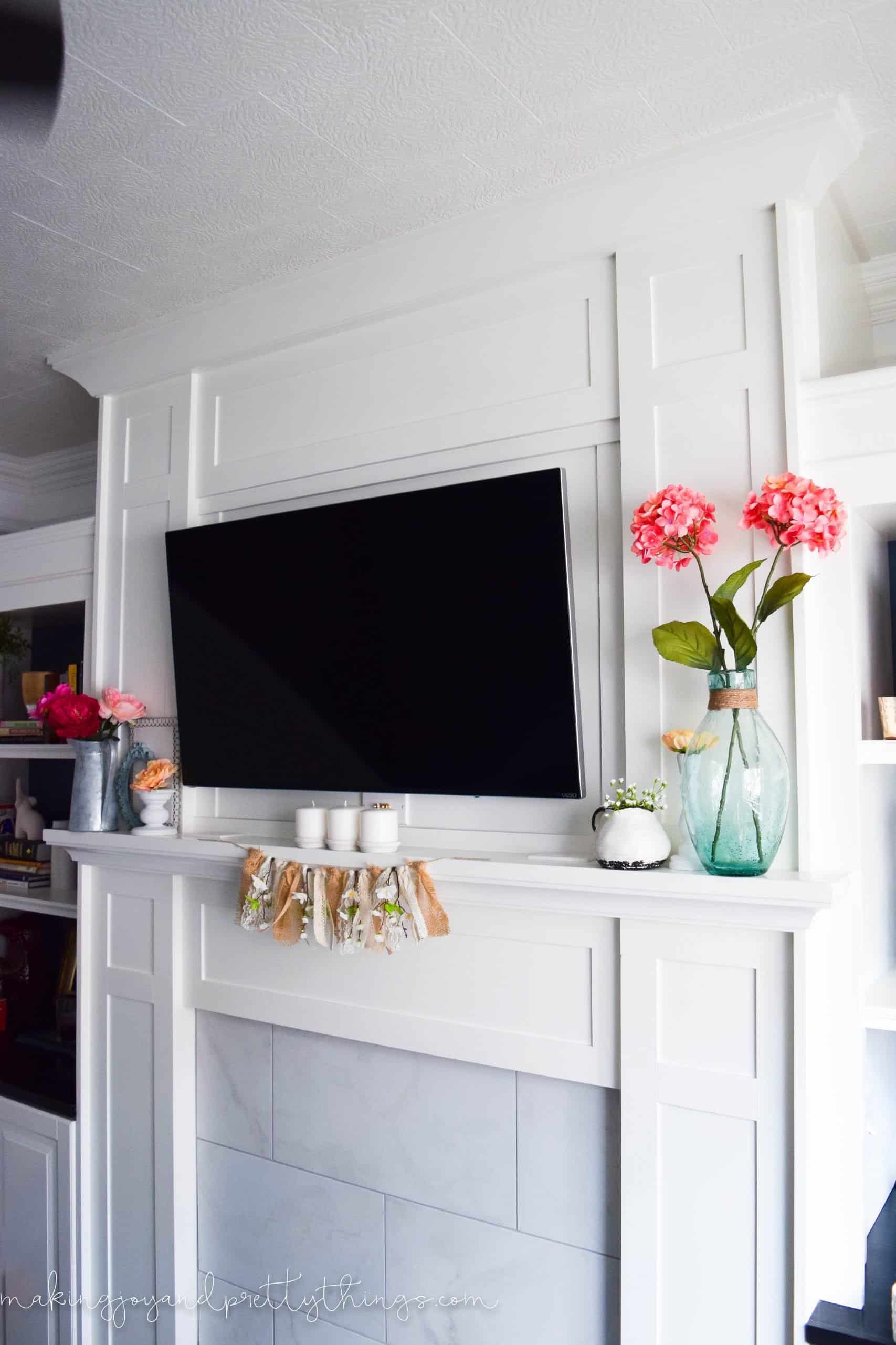 Another angle of our summer mantle decor - vases of bright pink faux flowers, pottery, and candles surround a wall-mounted flat screen TV above a white wood and marble mantle.