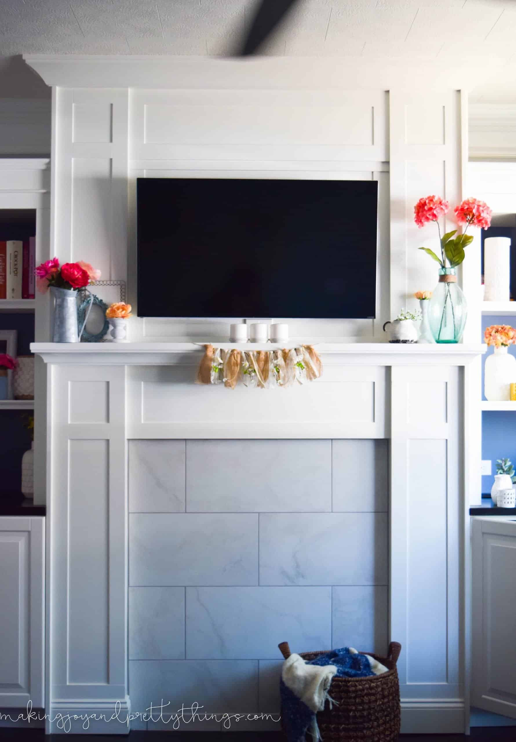 The completed summer mantle decorated with vases of faux flowers, a small peach blossom garland, decorated around a mounted flat screen tv.