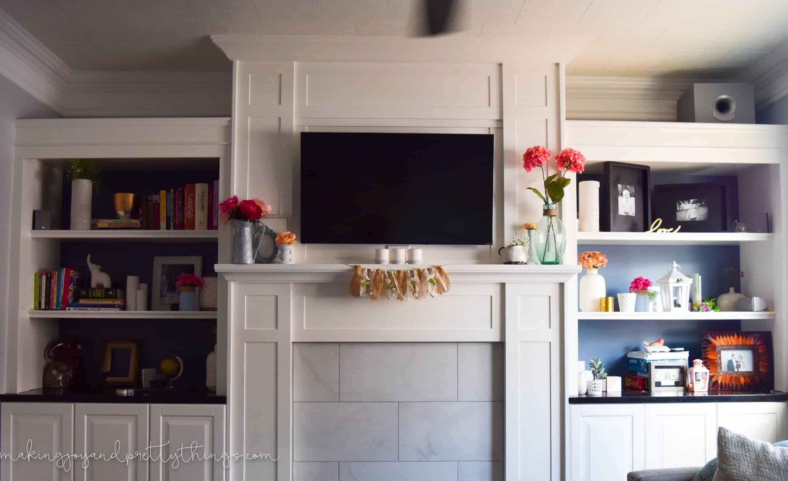 Our finished summer mantle decor on our living room mantle. The mantle stands between two built-in bookshelves filled with farmhouse decor. A large flatscreen TV is mounted on the mantle wall.