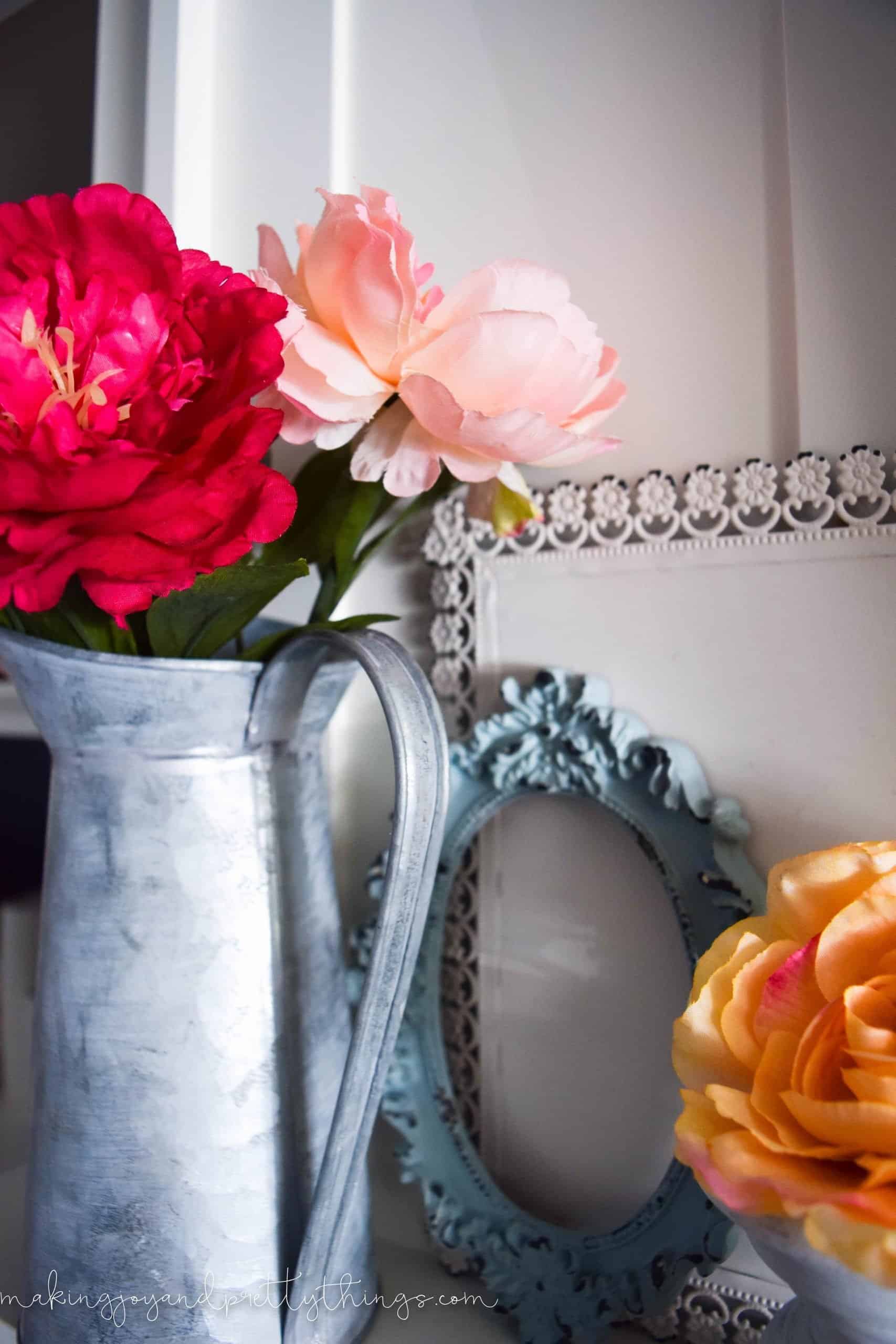 A galvanized metal vase filled with pink and red faux flower stems sits on the white mantle, next to two empty picture frames painted white and blue.
