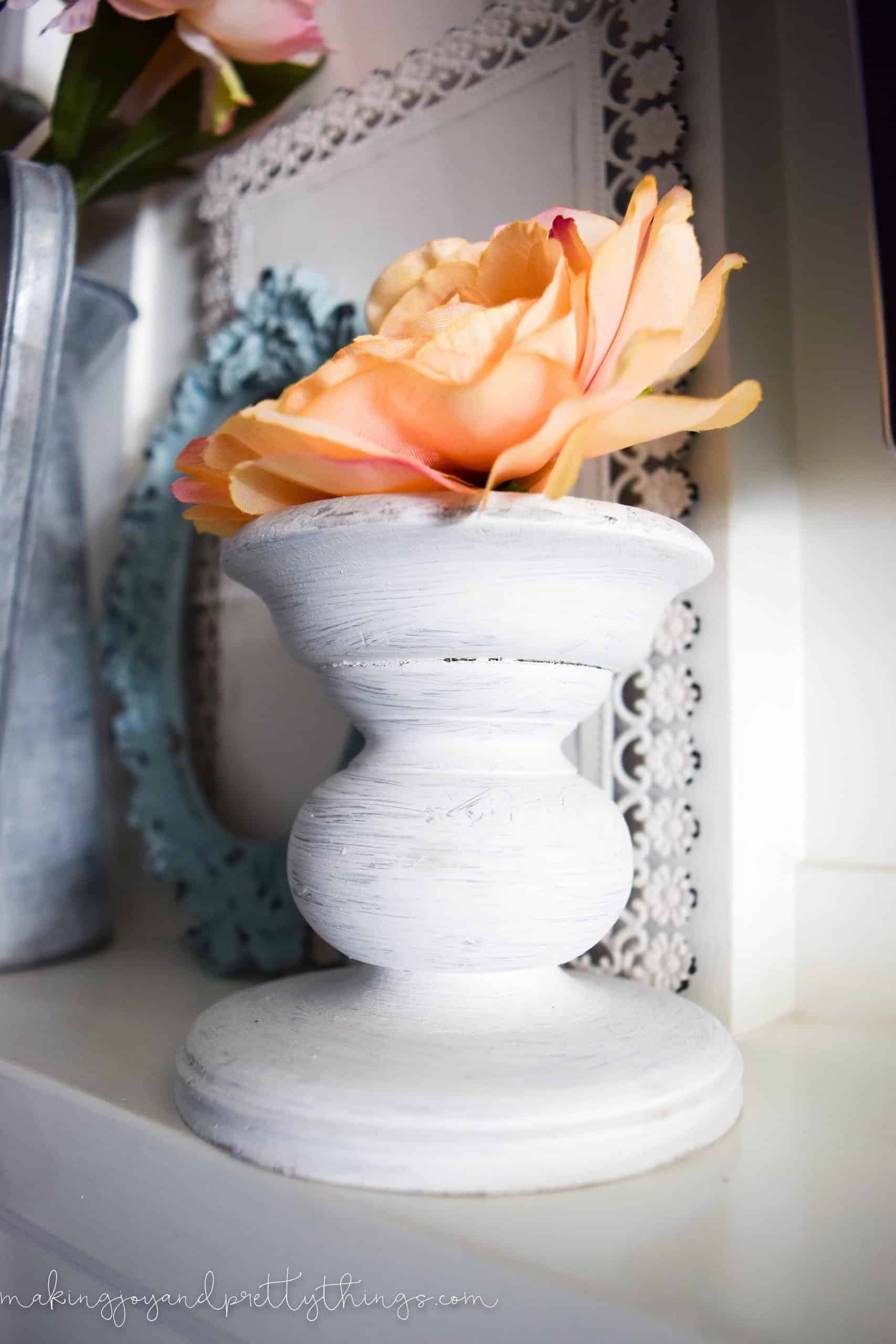 A rustic white painted candle stand holds a single light peach colored faux floral bloom.