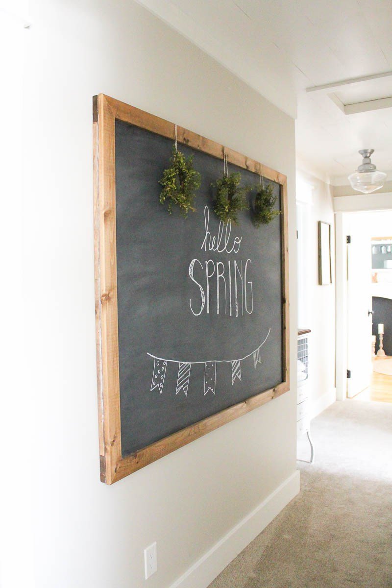 Large chalkboard with wooden frame in hallway with three small farmhouse wreaths hanging across the top with Hello Spring written on the chalkboard