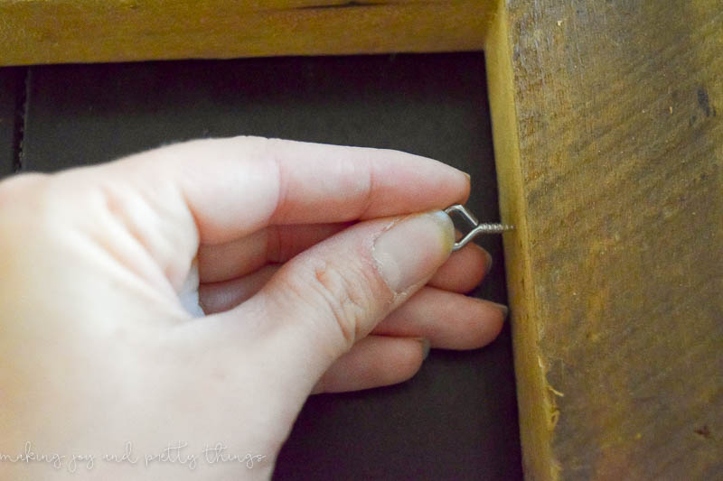 Image shows a woman's hand screwing a small picture eye hook into a rustic photo frame.