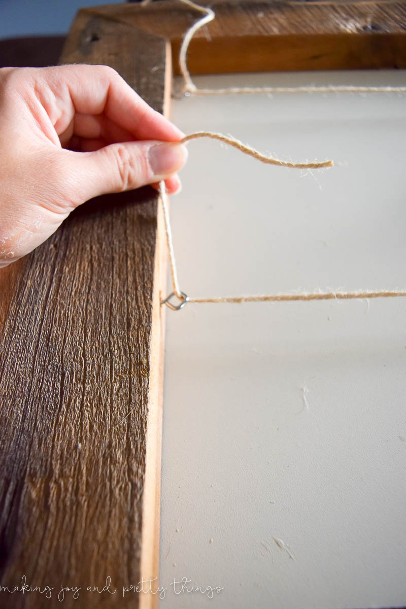 An image shows a woman's hand stringing a piece of twine between an eye hook that's attached to a wood frame. 