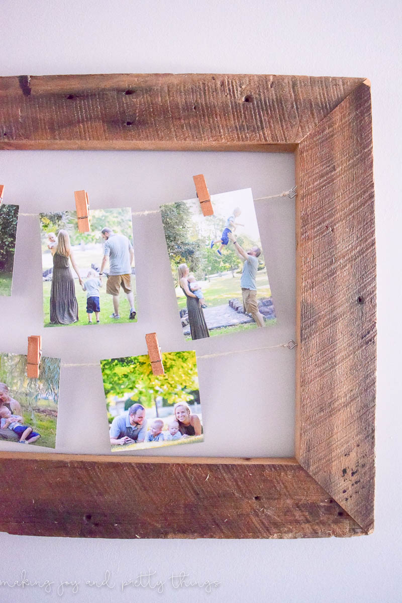 A DIY rustic picture frame made with reclaimed barnwood holds two strings of twine on eye hooks. Family pictures of a mom, dad, and little boy outside playing hang on the twine with clothespins.