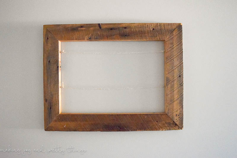 A rustic picture frame made with reclaimed barnwood hangs on a white wall. The picture frame is empty except for two strings of twine attached to eye hooks across the middle.