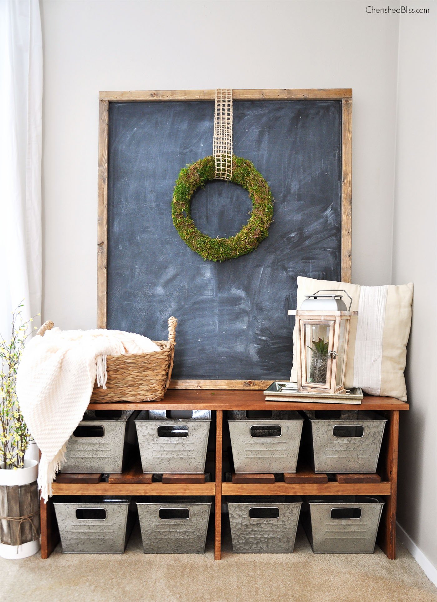 Large rustic oversized chalkboard with wooden frame with a moss wreath hanging in the middle leaning against a wall above a wooden bench with other farmhouse style decorations around it