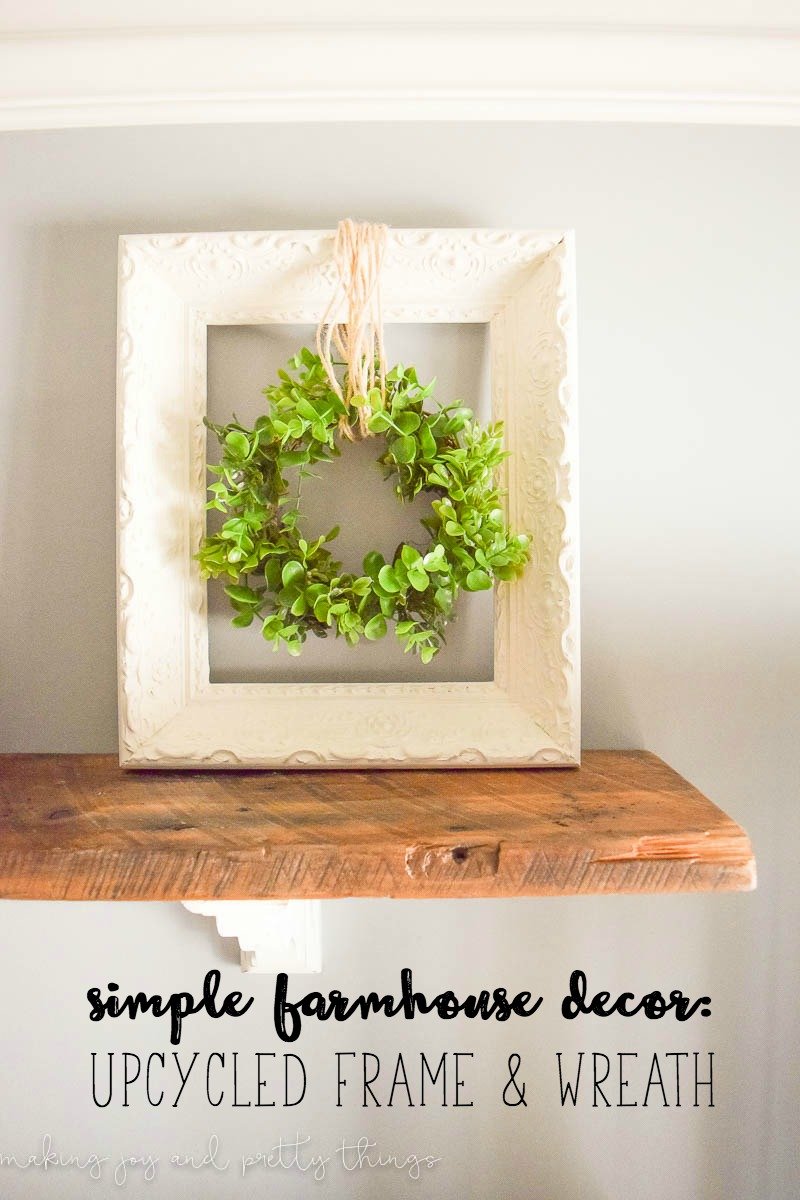 A cream-colored picture frame sits on a rustic wood shelf. A faux grapevine wreath hangs in the center of the picture frame from twine. This farmhouse style wreath hanger is perfect for rustic style decor.