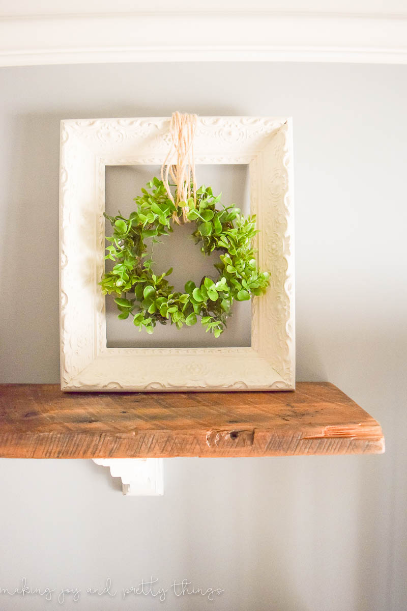 A farmhouse style greenery wreath hangs in a white picture frame on a rustic wood shelf.