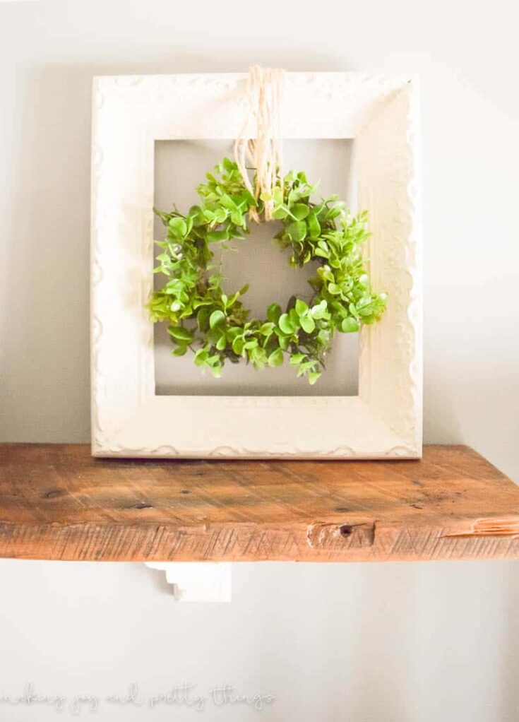 A farmhouse style wreath hanger sits on a wood shelf. A white picture frame holds a greenery wreath hanging from twine.