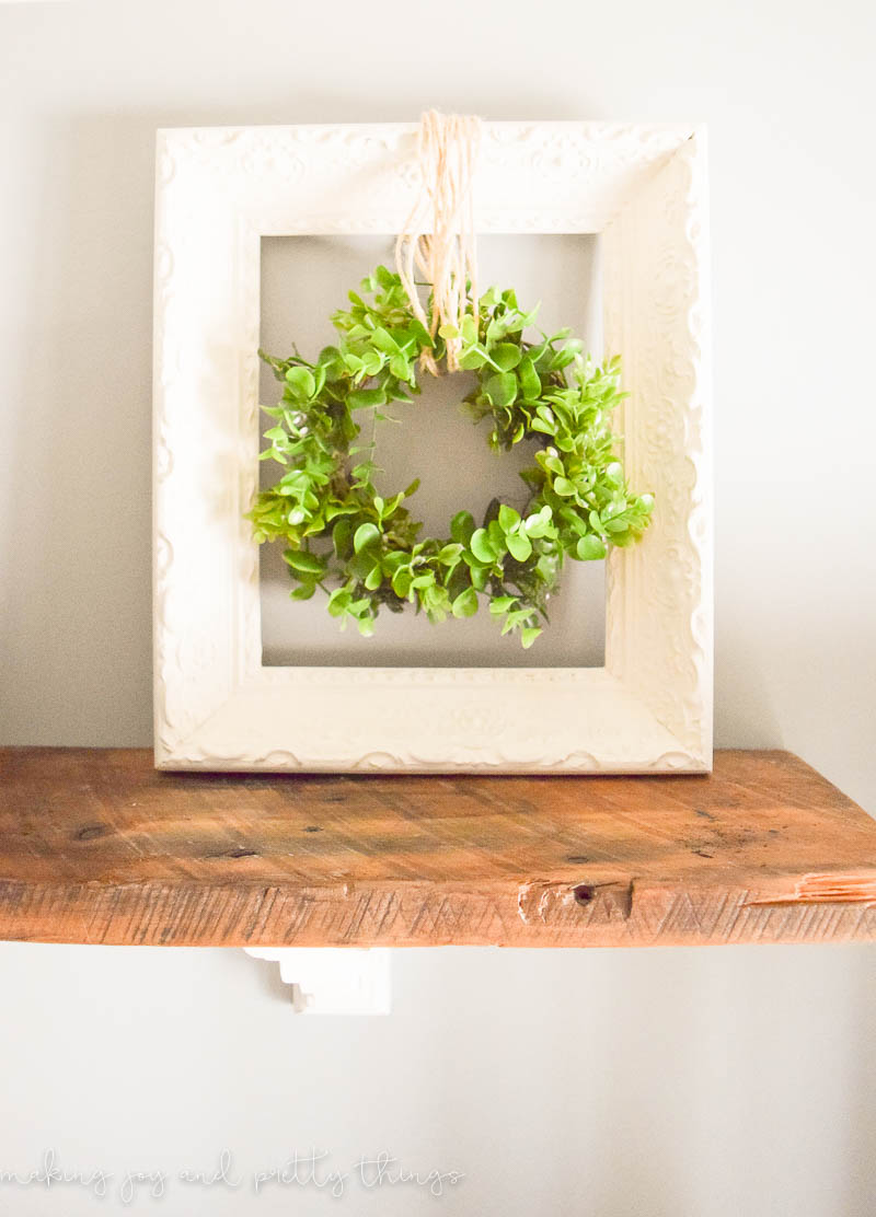 A cream-white painted picture frame sits on a rustic wood shelf against a white wall. In the center of the frame is a faux greenery wreath, hung with twine.