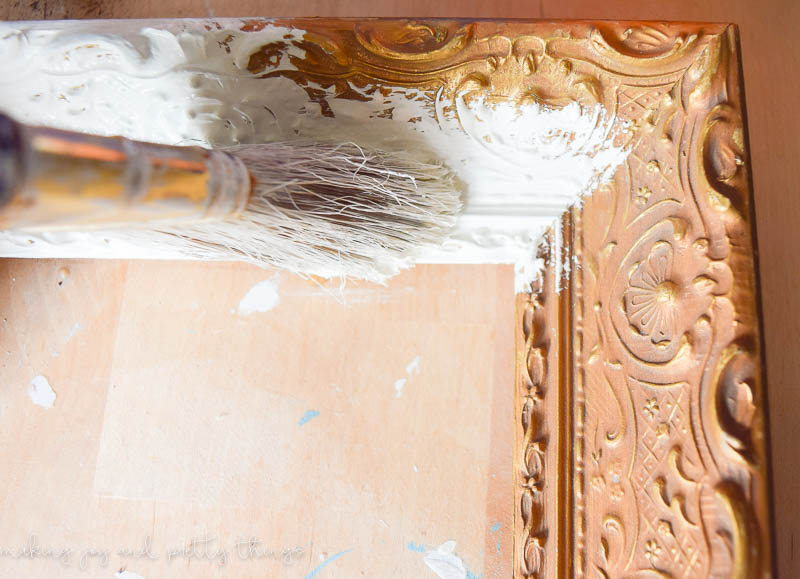 A close up look at the process of painting an old thrifted picture frame. A paint brush applies a linen white paint over the old, worn-out wood finish.