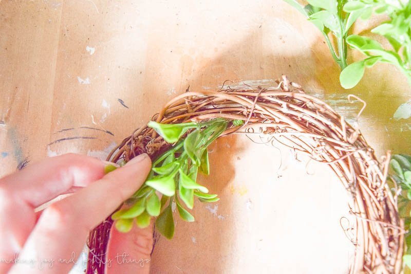 A woman's hang inserts faux greenery into a small grapevine wreath. 