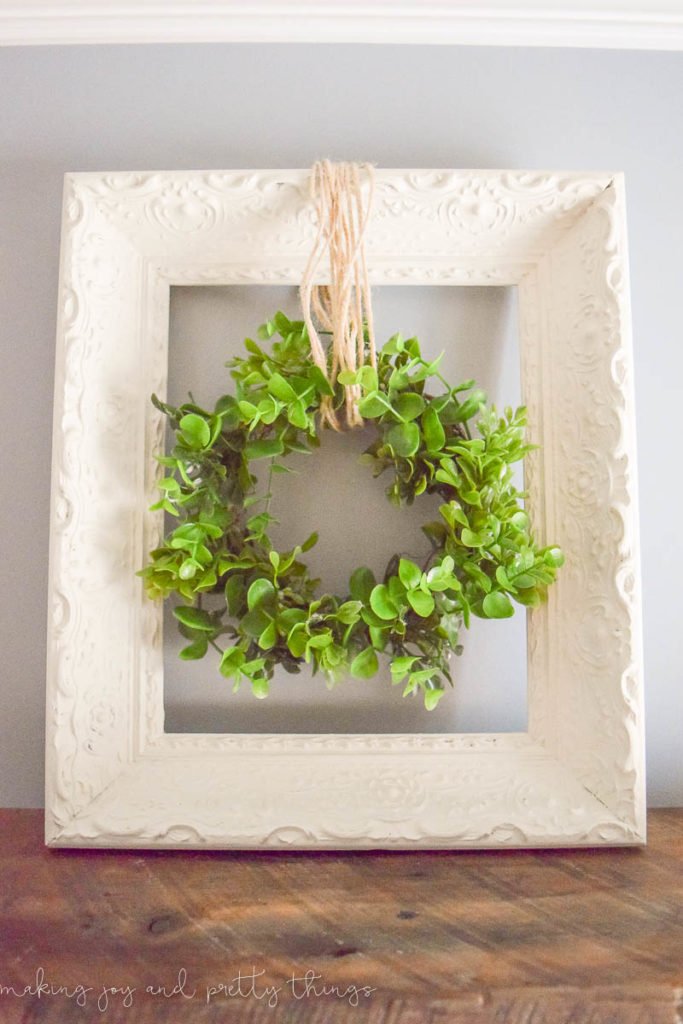 A small faux greenery wreath hangs from twine in the center of a white picture frame with an ornate carved pattern.