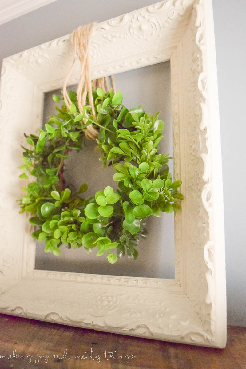 A faux greenery wreath hangs from twine inside an upcycled picture frame. The frame is painted a clean white linen color, with ornate detailing, and sits against a white wall.