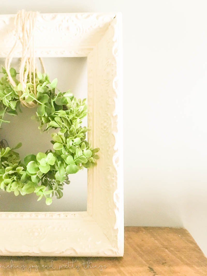 Image shows half of the DIY wreath hanger made with an upcycled wood frame that's been painted a crisp, clean white. A greenery wreath hangs in the center of the frame from looped twine.