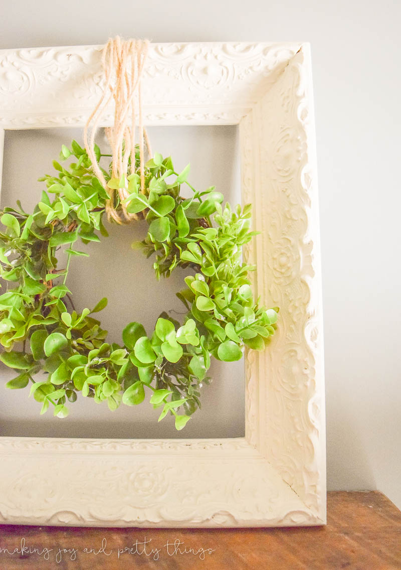 A closer look at the recycled picture frame wreath hanger. The ornate details of the wood picture frame can be seen though the cream white paint. A faux greenery wreath hangs from twine.