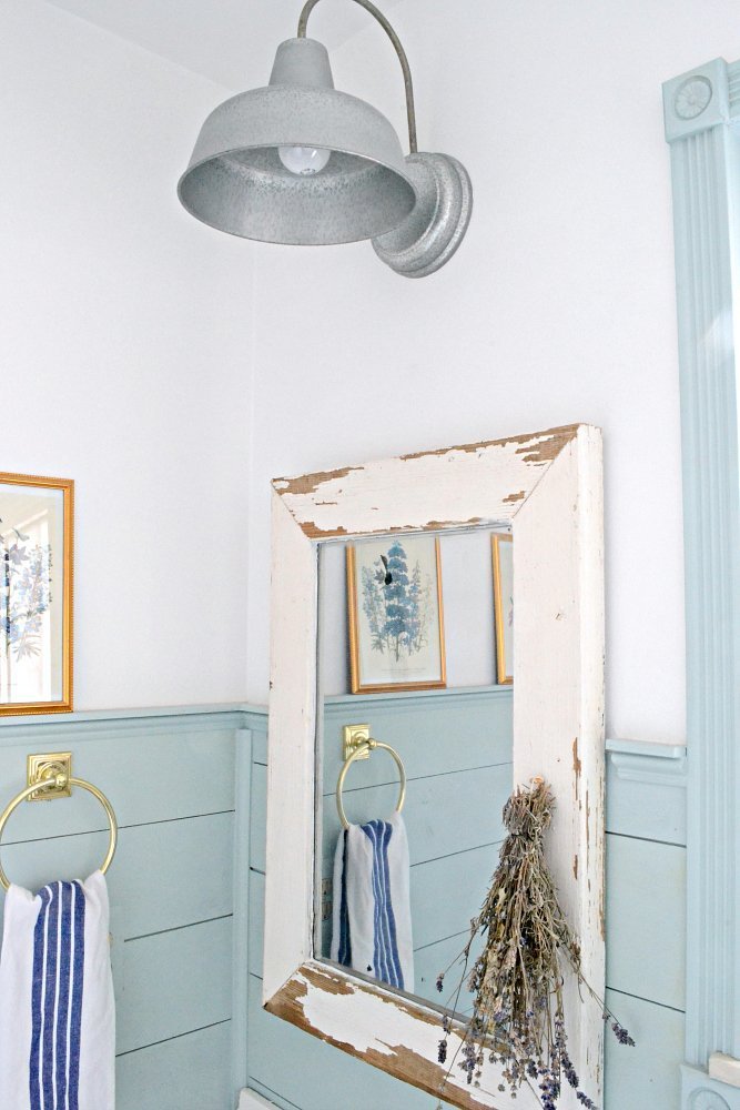 Farmhouse mirror with white chippy paint frame made from a salvaged window hanging on the wall in a bathroom with dried lavender hanging upside down on the frame and towel rack on the wall beside it 