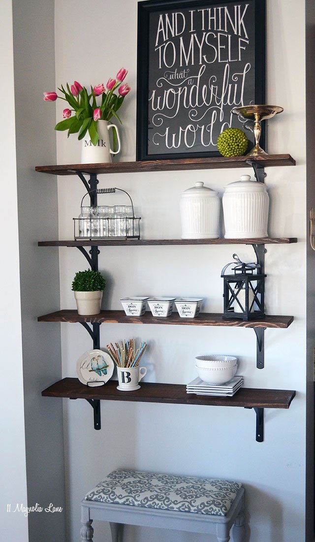 Wooden open shelves stained with dark wood stain hanging on the wall with metal brackets decorated with faux plants, frames, bowls, candles, frame with quote, jars and canisters 