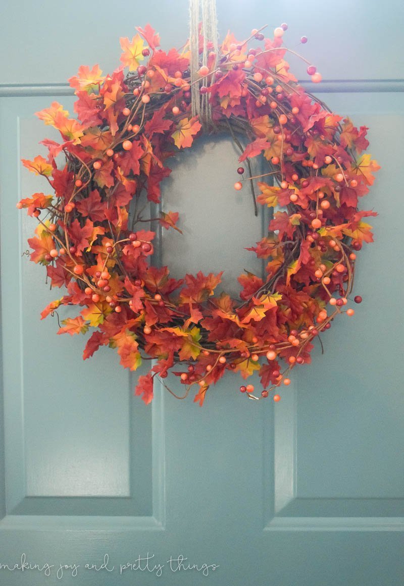 A completed fall-inspired leaf wreath hangs against a green front door. The wreath is filled with vibrant red, orange, and yellow leaves and branches of faux berries, and hangs from loops of twine.