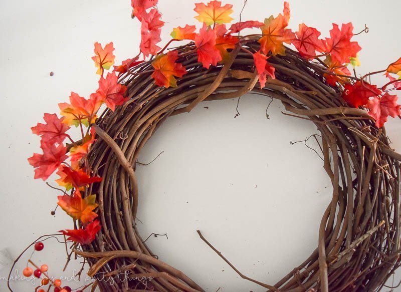 A grapevine wreath with a strand of fall leaf garland wrapped half way around. The leaves are a mix of bright orange and yellow.