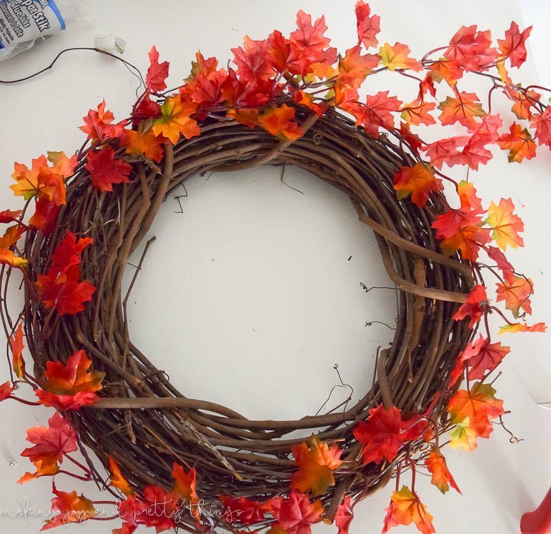 A grapevine wreath wrapped in strands of leaf garland, with bright orange, red and yellow leaves.