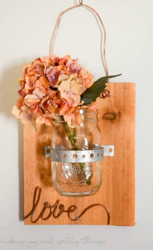 Mason jar with faux flower in it attached to piece of scrap wood with love written in wire hanging on a wall