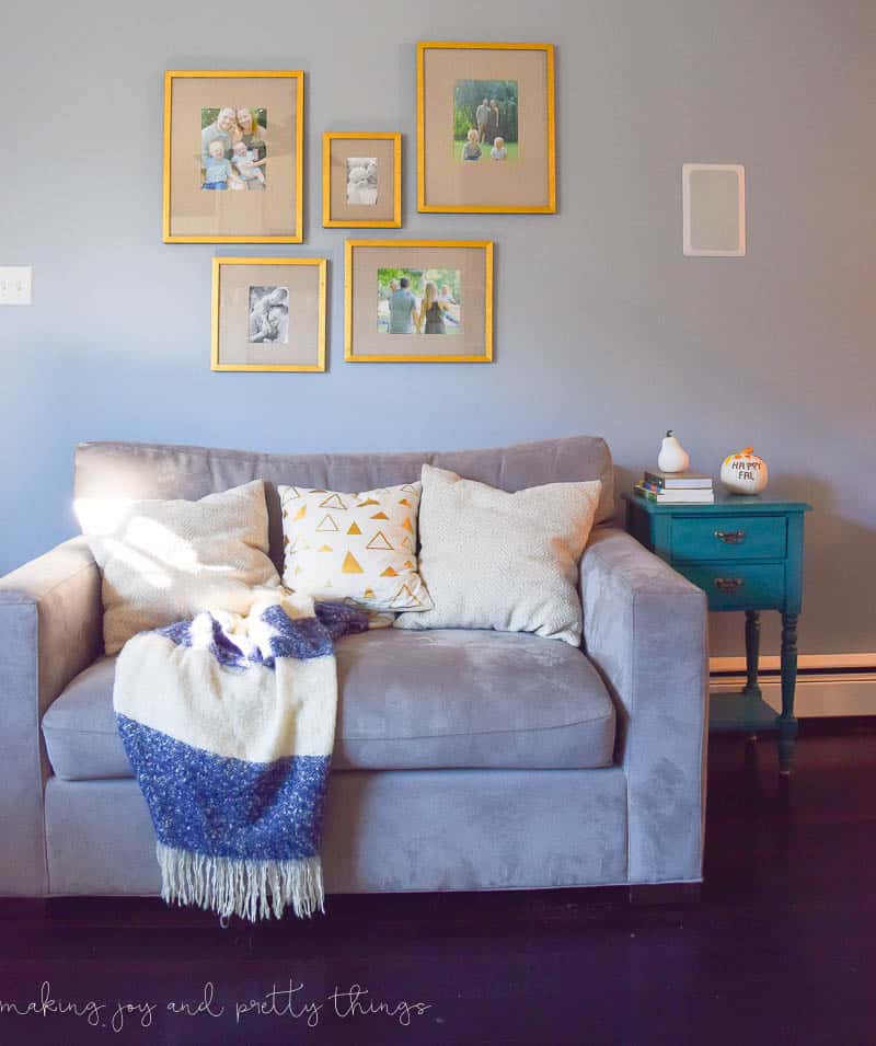 An oversized chair decorated with blankets and throw pillows sits under a gallery wall. Five photos are framed in different sized gold frames hanging on the gray wall.