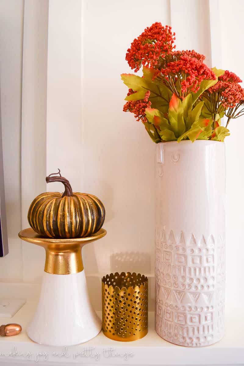 A white vase filled with fall themed flowers, a gold decorative pumpkin on a gold and white pedestal stand, and a gold candle holder sit on a white fireplace mantle.