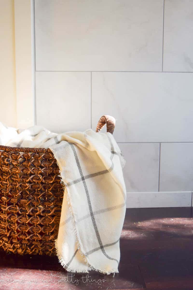 A woven wood basket  holds a white plaid throw blanket, on the floor in front of a white marble fireplace mantle.