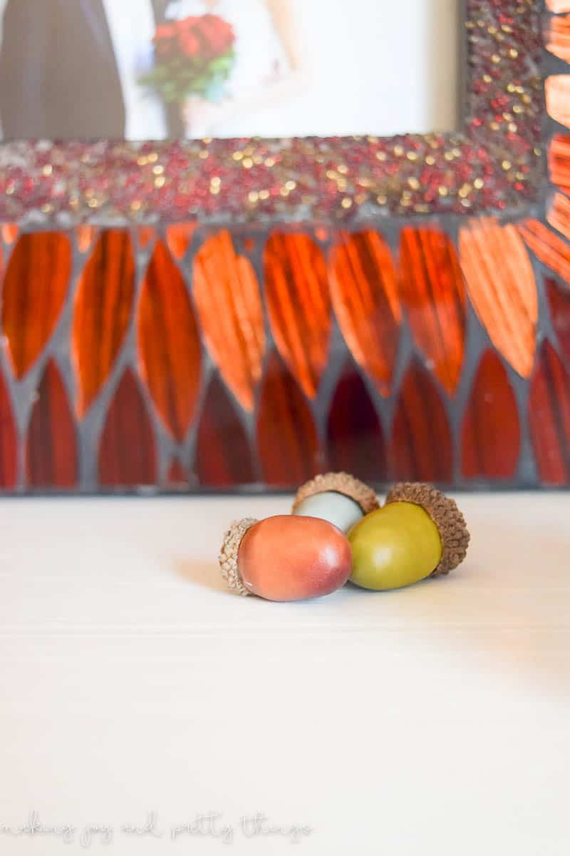 A close up look at three mini acorns sitting on a shelf in front of a bright orange picture frame. The tiny acorns are orange, green, and blue.