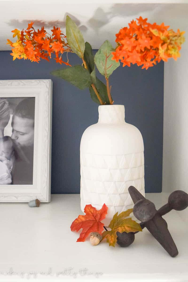 A white ceramic vase sits on a white shelf and is filled with faux flowers - bright orange and yellow florals with green leaves.