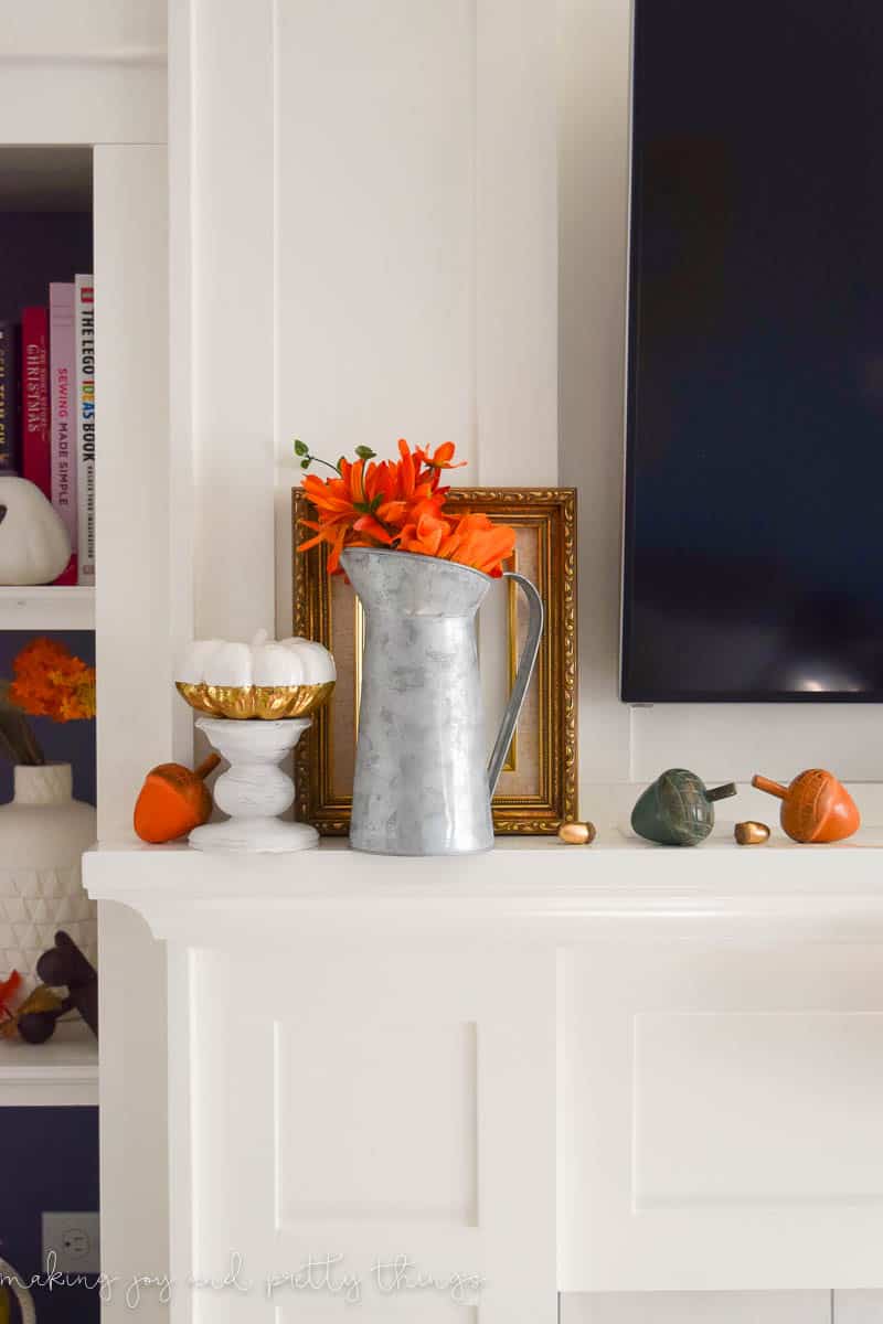 A closer look at the left side of the living room mantel, decorated with a tin watering can vase filled with orange flowers, colorful decorative acorns, and a white and gold painted pumpkin.