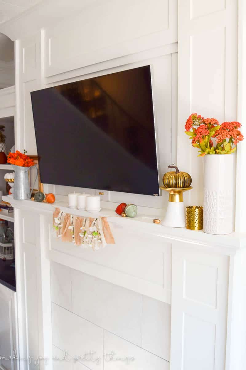 A different angle of our living room mantel decorated with Fall decor. Vases of fall flowers, candles, faux pumpkins, and a floral banner hangs on the mantle. A flat screen tv is mounted on the wall.