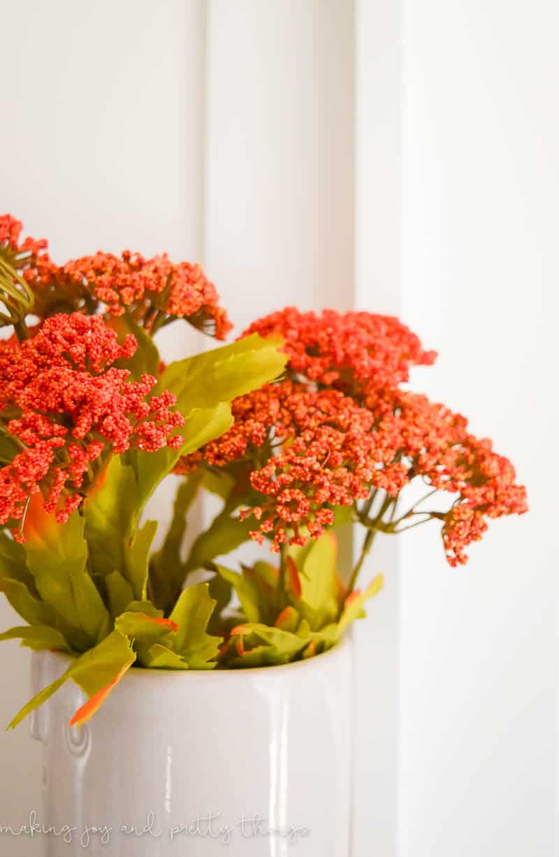 A close up look at the faux fall flowers in a white vase. Small orange beaded flower heads and light green leaves. 