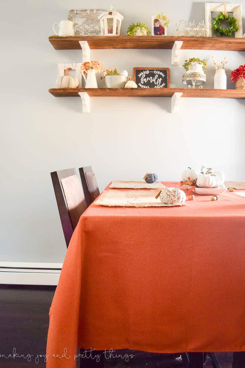 Another look at our Fall dining room scene - a dining table covered with a burnt orange table cloth and colorful fall centerpiece. Floating shelves are filled with traditional fall decor pieces - vases and faux florals and pumpkins, picture frames and more.