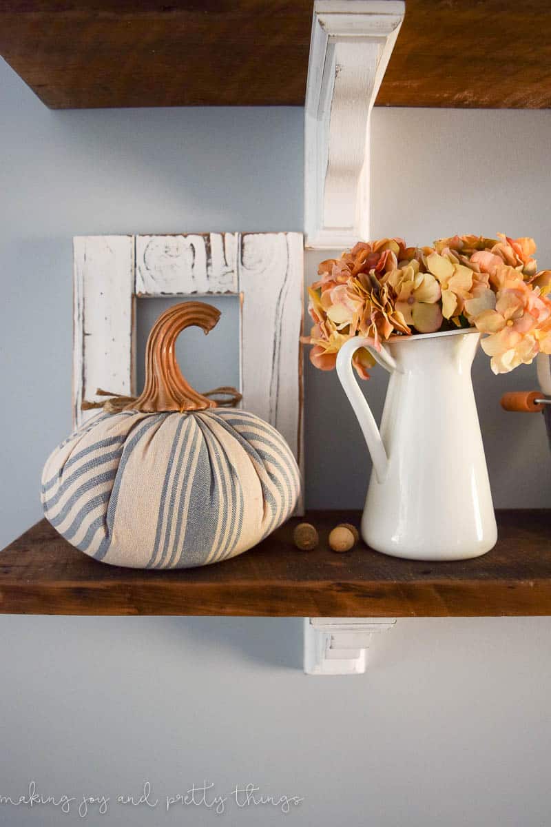 A white milk can vase is filled with faux flowers, and sits next to a blue and white stripped fabric pumpkin with a wooden stem.