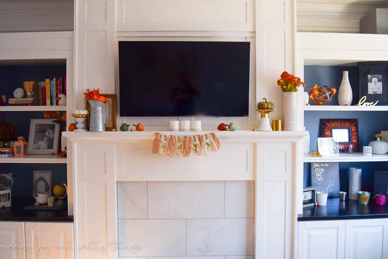 A white living room mantel, decorated with traditional fall decor. Vases filled with fall-colored flowers, candles, decorative pumpkins and acorns line the mantel. On either side are built-in bookcases.