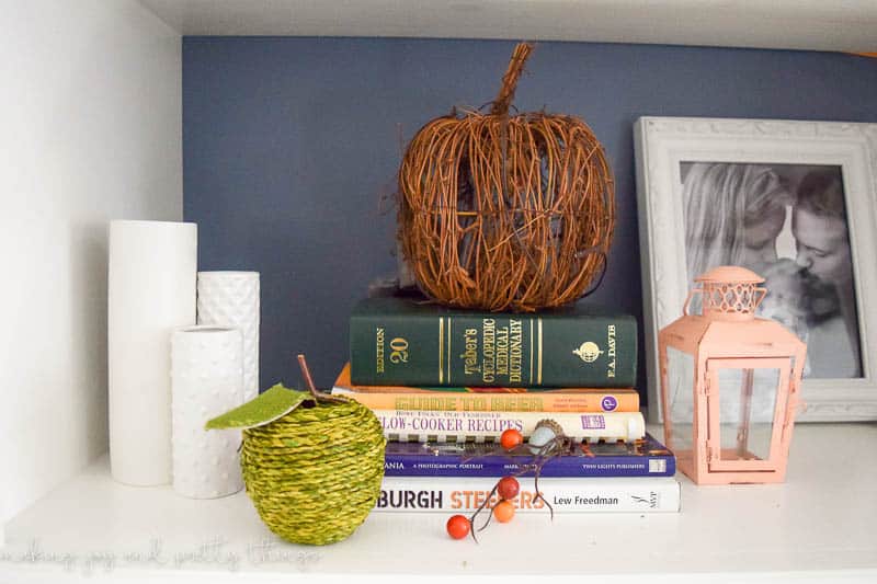 Another shelf of our living room built-ins decorated with a stack of books, a framed picture, small peach-colored lantern, three white pillar candles, a wood pumpkin, mini faux acorns, and a green apple made from woven rope.