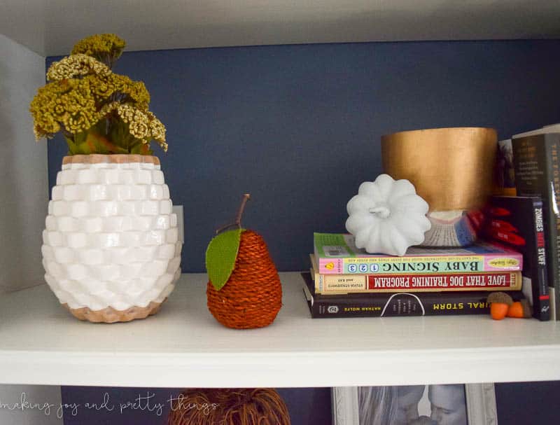 Another shelf of our living room built-ins decorated with stacks of books, mini faux pumpkins, a small pear made with woven rope, and a white and gold vase holding textured florals.