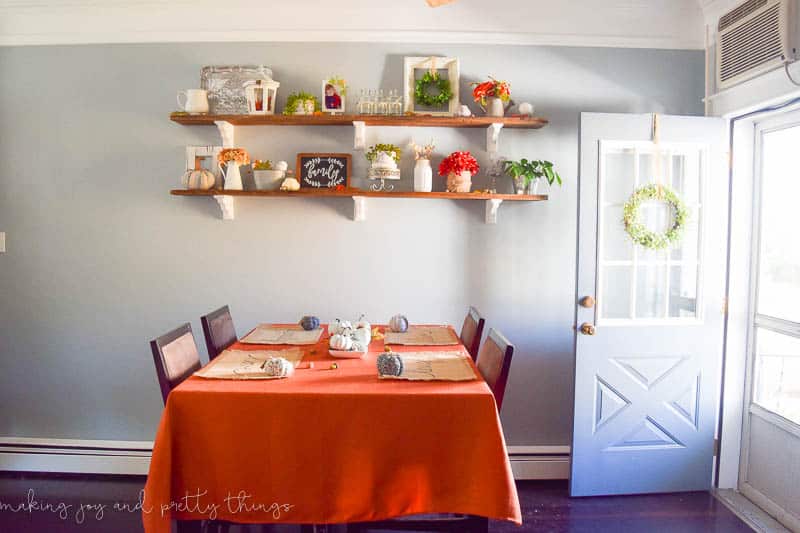 A small dining room scene is arranged with a table with four chairs and place settings, covered with a burnt orange table cloth. Above the dining table are wall mounted floating shelves filled with Fall decor pieces - vases, flowers, picture frames and more.
